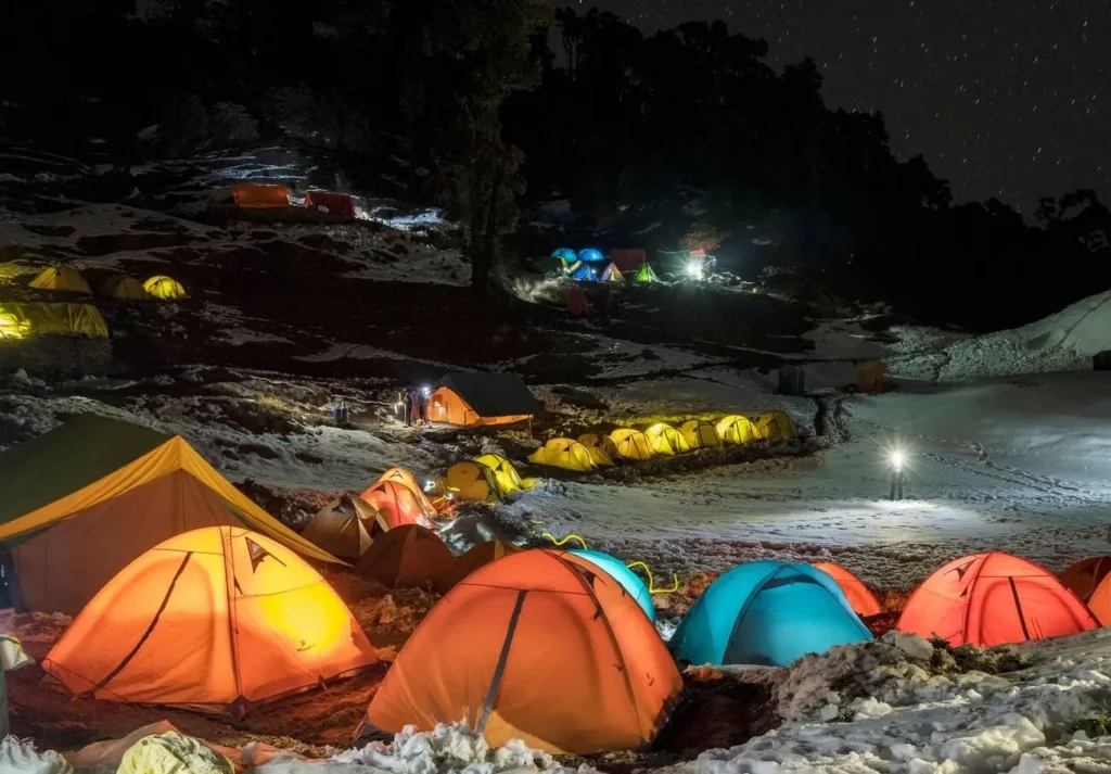 Riverside camping site near Jibhi, Himachal Pradesh, with illuminated tents under a clear, starry sky.